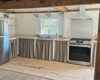 May include: A kitchen with light wood floors and exposed wooden beams. The kitchen features a stainless steel refrigerator, a sink, and a stainless steel oven. Beige curtains hang below the countertops. The walls are painted white.