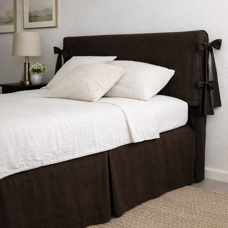 May include: A dark brown bed headboard with matching bed skirt. The headboard has decorative bows on the sides. White pillows and bedding are on the bed. The room has a neutral color palette.