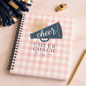 Könnte beinhalten: Ein spiralgebundenes Notizbuch mit einem rosa-weiß karierten Muster. Das Cover zeigt ein marineblaues Megaphon mit dem Wort "cheer" und dem Text "CHEER COACH Plan. Lead. Inspire." Ein goldener Stift, eine Pfeife und marineblaue und goldene Pompons sind ebenfalls sichtbar.