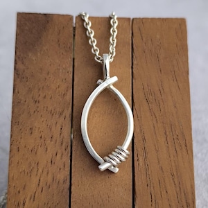 May include: A silver-colored pendant necklace featuring an oval design with a wrapped wire accent. The pendant hangs from a delicate chain and is displayed on a wooden block. The background is a soft gray.