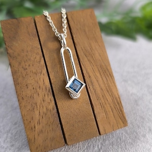 May include: A silver pendant necklace with a blue gemstone set in a silver frame. The pendant is shaped like a long, thin rectangle with a rounded top and bottom. The necklace is on a wooden display stand.