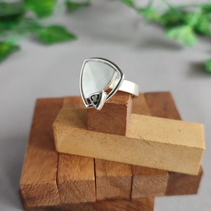 Könnte beinhalten: Ein silberner Ring mit einem dreieckigen Stein, der in der Mitte eingesetzt ist. Der Stein ist hellgrau mit einem dunkelgrünen Zentrum. Der Ring liegt auf einem Stapel Holzblöcken.