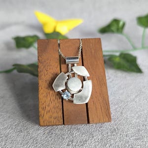 Puede incluir: Un collar con un colgante de plata de diseño geométrico, con un centro circular y una piedra preciosa azul. El colgante se muestra en un soporte de madera.