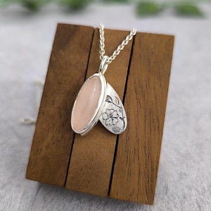 May include: A silver pendant necklace with an oval, light pink stone. The pendant has a floral design on one side. The necklace is displayed on a wooden stand against a neutral background.