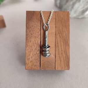 Op de afbeelding: Een zilveren ketting met een hanger met een stapel gouden en zilveren ringen die aan een gehamerde zilveren staaf zijn bevestigd. De ketting wordt gepresenteerd op een houten standaard.