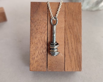 May include: A silver necklace with a pendant featuring a stack of gold and silver rings attached to a hammered silver bar. The necklace is displayed on a wooden stand.