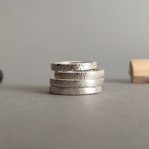 May include: A stack of five silver rings with different textured finishes. The rings are stacked on top of each other, with the top ring having a hammered finish, the second ring having a brushed finish, the third ring having a hammered finish, the fourth ring having a brushed finish, and the bottom ring having a hammered finish.