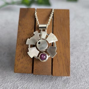 Op de afbeelding: Een zilveren ketting met een geometrische hanger met een klein zwart steentje en een groter roze steentje. De hanger ligt op een houten standaard.