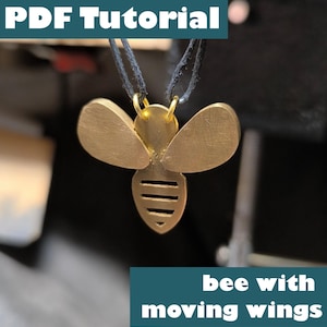 Può includere: Un ciondolo a forma di ape color oro con ali mobili, sospeso a un cordino nero. Il design dell'ape presenta un corpo e ali dettagliati. Il testo sull'immagine recita "PDF Tutorial" e "bee with moving wings".