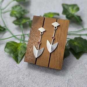 Peut inclure: Une paire de boucles d'oreilles en argent avec des accents de pierres précieuses rouges. Les boucles d'oreilles présentent un design géométrique avec une forme de flèche pointue en bas.