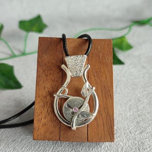 Könnte beinhalten: Eine silberne Halskette mit einem zarten Blumenmuster und einem rosafarbenen Edelstein. Der Anhänger ist an einer schwarzen Schnur befestigt und auf einem Holzständer ausgestellt.