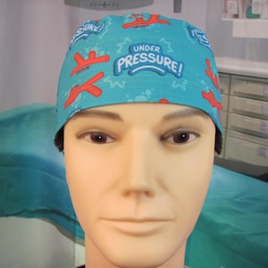 Peut inclure: Un bonnet de chirurgien bleu turquoise avec des accents rouges et blancs. Le bonnet porte l'inscription "Under Pressure !" imprimée dessus.