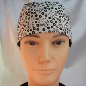 May include: A white scrub hat with a black and yellow leopard print and the word "SIMBA" repeated throughout the design. The hat features a cartoon image of a lion cub.