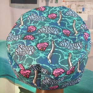 May include: A blue and green patterned surgical cap with the words "Neuro Surgery" repeated throughout the design. The cap features a stylized brain and spinal cord design.