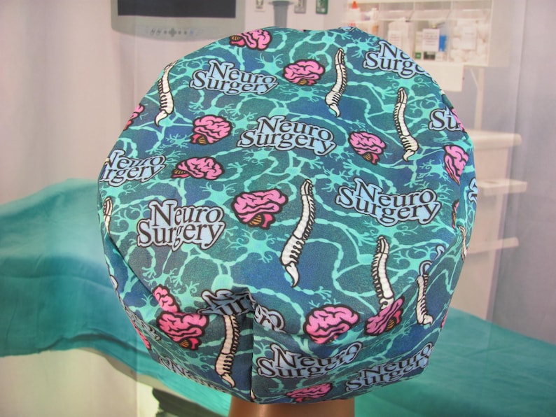 May include: A blue and green patterned surgical cap with the words "Neuro Surgery" repeated throughout the design. The cap features a stylized brain and spinal cord design.