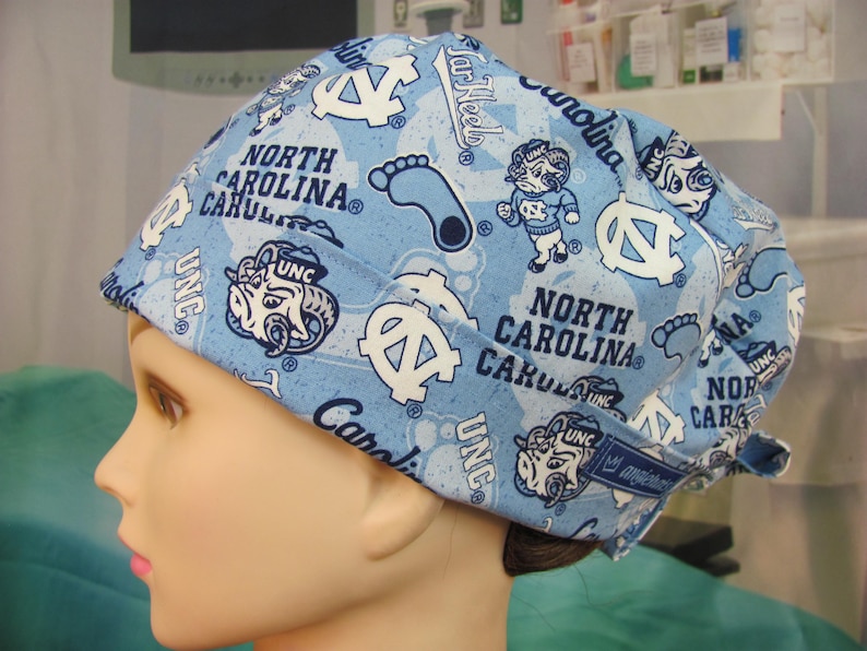 May include: A light blue scrub cap with a repeating pattern of UNC logos, the words "North Carolina," and the UNC mascot. The cap is made of a soft fabric and is designed for medical use.