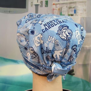 May include: A light blue surgical scrub cap featuring a repeating pattern of UNC logos, the words "North Carolina", and other university-related graphics in white. The cap is tied at the back.