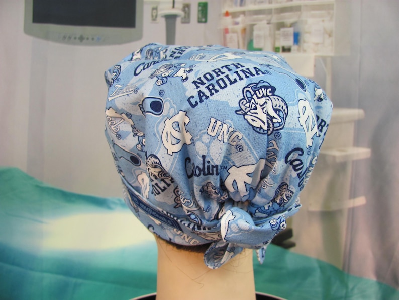May include: A light blue surgical scrub cap featuring a repeating pattern of UNC logos, the words "North Carolina", and other university-related graphics in white. The cap is tied at the back.