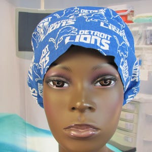 May include: Blue surgical cap with a white print of the Detroit Lions logo. The cap is worn by a person with dark skin.