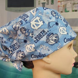 May include: A light blue scrub cap with a repeating pattern of white logos, text, and cartoon figures. The cap features the words "North Carolina", "UNC", and "Tar Heels". The cap is tied at the back.