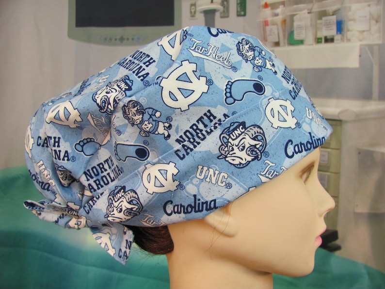 May include: A light blue scrub cap with a repeating pattern of white logos, text, and cartoon figures. The cap features the words "North Carolina", "UNC", and "Tar Heels". The cap is tied at the back.