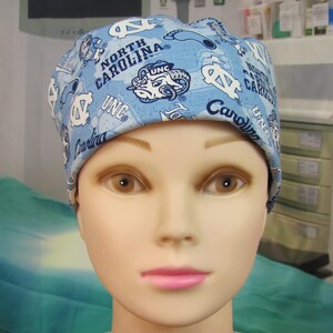 May include: A light blue scrub cap with the text "North Carolina" and "UNC" and the university's ram mascot. The cap is patterned with university-related graphics and logos. The cap is worn by a mannequin.