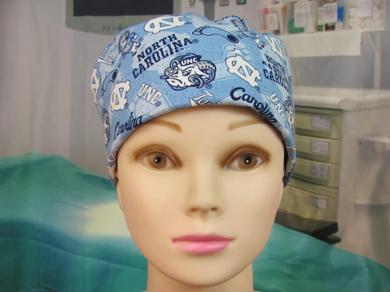 May include: A light blue scrub cap with the text "North Carolina" and "UNC" and the university's ram mascot. The cap is patterned with university-related graphics and logos. The cap is worn by a mannequin.