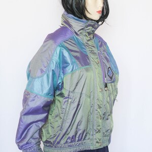 Pu&ograve; includere: Una giacca iridescente di colore viola, verde e blu con chiusura a zip e bottoni a pressione. La giacca ha un colletto alto e una vita con coulisse.
