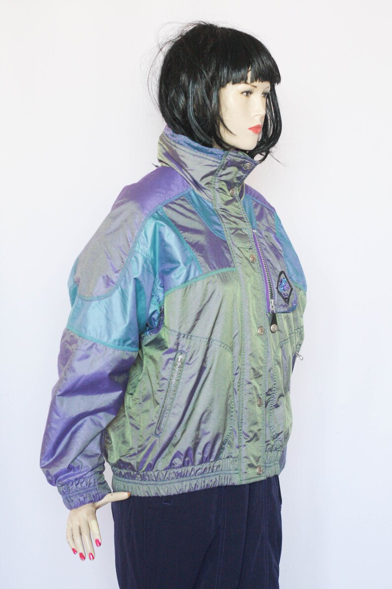 Pu&ograve; includere: Una giacca iridescente di colore viola, verde e blu con chiusura a zip e bottoni a pressione. La giacca ha un colletto alto e una vita con coulisse.