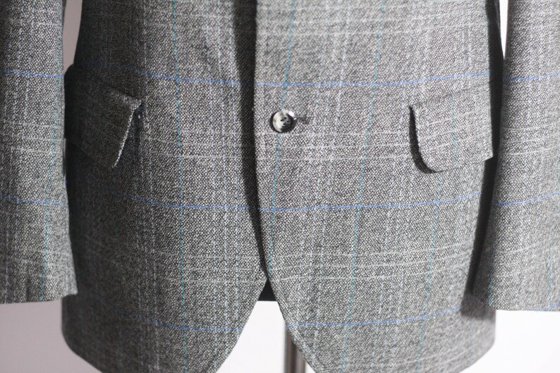 Pu&ograve; includere: Un blazer grigio con un motivo a quadri blu e bianchi. Il blazer ha una chiusura con un solo bottone e due tasche a lembo.
