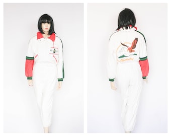 May include: White ski suit with red and green accents. The suit features a zipper closure, a high collar, and a logo that says "HED" with a mountain graphic. The back of the suit features a graphic of a bird flying over a mountain range.