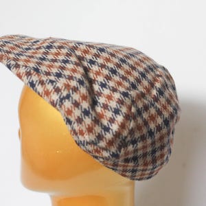 May include: A brown, navy, and white houndstooth pattern newsboy cap. The cap is made of a textured fabric and has a rounded crown and a small, stiff brim. The cap is displayed on a mannequin head.