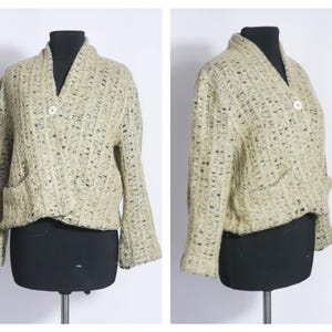 May include: A cream-colored knitted cardigan with a shawl collar, long sleeves, and two pockets. The cardigan has a button closure and a textured knit pattern.