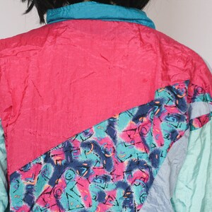 Puede incluir: Una chaqueta cortavientos de color rosa brillante, azul verdoso y azul con un patr&oacute;n geom&eacute;trico. La chaqueta tiene una cremallera y un cuello de pie.