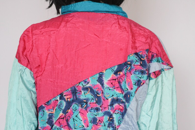 Puede incluir: Una chaqueta cortavientos de color rosa brillante, azul verdoso y azul con un patr&oacute;n geom&eacute;trico. La chaqueta tiene una cremallera y un cuello de pie.