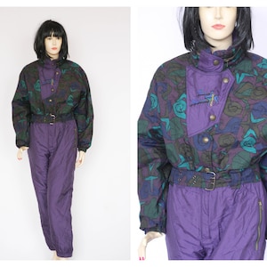 May include: Purple ski suit with a black, green, and blue abstract print. The suit has a zipper closure, a belt, and a hood.