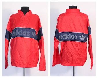 Może przedstawiać: Czerwona kurtka wiatrówka Adidas z niebieskimi i białymi lamówkami i logo Adidas z przodu i z tyłu.