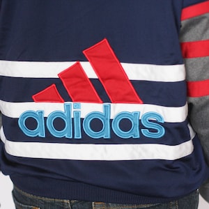 Peut inclure: Une veste ray&eacute;e bleu et blanche avec des accents rouges et gris. La veste pr&eacute;sente un grand logo Adidas brod&eacute; en rouge, blanc et bleu.