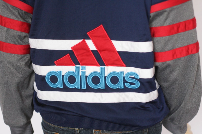 Peut inclure: Une veste ray&eacute;e bleu et blanche avec des accents rouges et gris. La veste pr&eacute;sente un grand logo Adidas brod&eacute; en rouge, blanc et bleu.