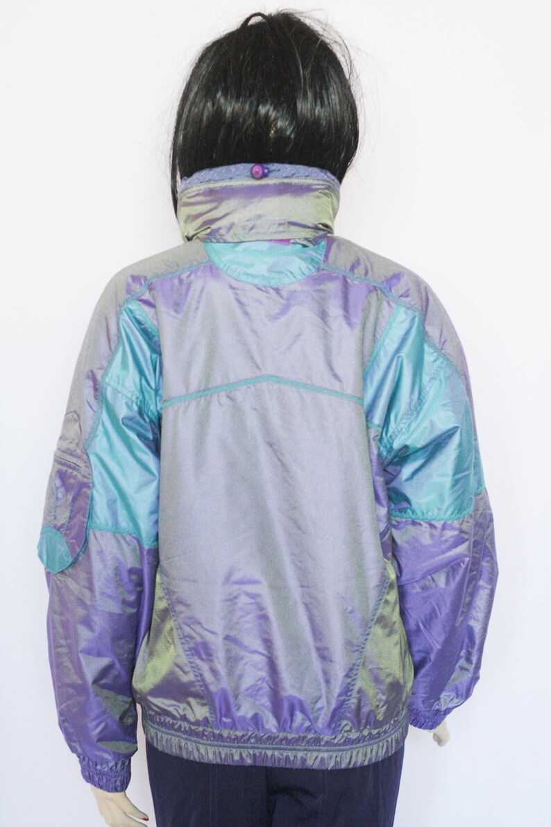 Pu&ograve; includere: Una giacca iridescente viola e turchese con chiusura a zip. La giacca ha un colletto alto e un cordoncino in vita.