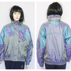 Pu&ograve; includere: Una giacca iridescente di colore viola, turchese e argento con chiusura a zip e un bottone a pressione sul collo. La giacca ha una tasca con zip sul lato sinistro.