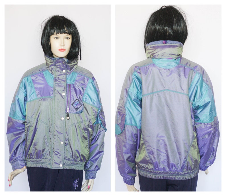 Pu&ograve; includere: Una giacca iridescente di colore viola, turchese e argento con chiusura a zip e un bottone a pressione sul collo. La giacca ha una tasca con zip sul lato sinistro.
