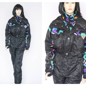 Peut inclure: Combinaison de ski noire avec un motif géométrique coloré. La combinaison a un col montant et une fermeture éclair.