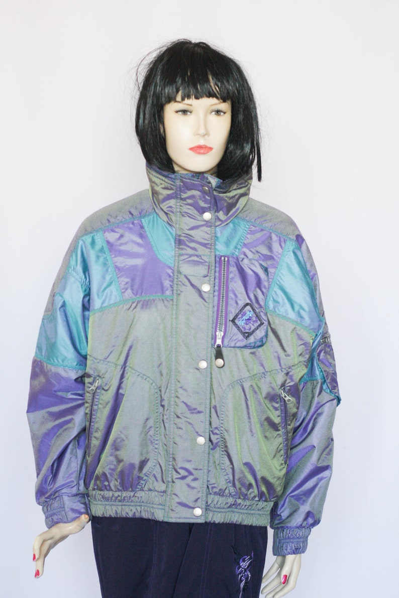 Pu&ograve; includere: Una giacca blu e viola iridescente con chiusura a zip e bottoni a pressione. La giacca ha un colletto alto e una vita con coulisse.