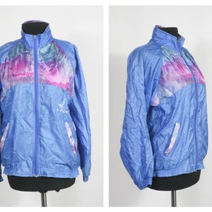 Puede incluir: Una chaqueta cortavientos azul con un estampado abstracto multicolor en la parte delantera y trasera. La chaqueta tiene cierre de cremallera y dos bolsillos.