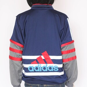 Peut inclure: Une veste Adidas bleu marine avec des rayures rouges et blanches et un grand logo Adidas au dos. La veste a des manches grises avec des rayures rouges.