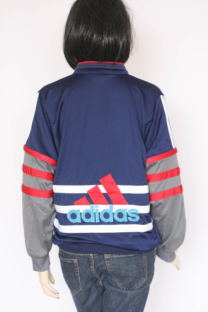 Peut inclure: Une veste Adidas bleu marine avec des rayures rouges et blanches et un grand logo Adidas au dos. La veste a des manches grises avec des rayures rouges.