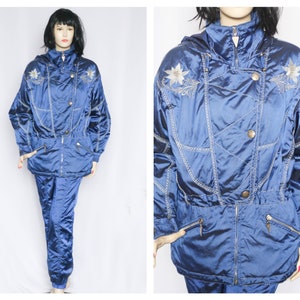 Può includere: Una tuta da sci blu con fiori bianchi ricamati. La giacca ha una cerniera lampo e un cappuccio. I pantaloni hanno una cintura elastica.