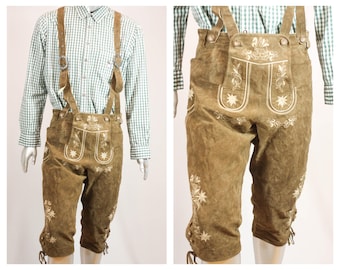 May include: Olive green suede leather lederhosen featuring adjustable suspenders, a front drop-front flap with white floral embroidery, and lace-up details at the leg cuffs. The garment is worn over a green and white checkered long-sleeve button-down shirt.