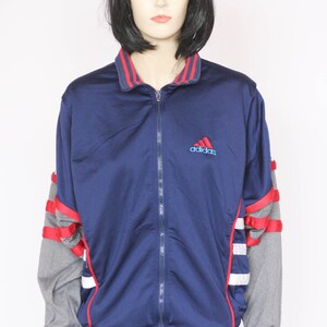 Peut inclure: Une veste zipp&eacute;e bleu marine avec des rayures rouges et blanches sur les manches. La veste a un panneau gris sous les bras et le logo Adidas sur la poitrine.